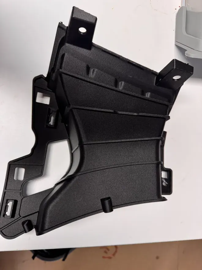 Pièce technique noire imprimée en 3D pour une carrosserie automobile, vue de trois quarts avec ses nervures et ses trous de fixation