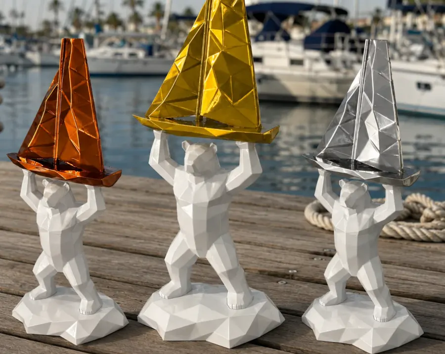 Trois trophées imprimés en 3D alignés sur un ponton : des ours blancs à facettes portant chacun un voilier bronze, or ou argent, avec un port de plaisance en arrière-plan